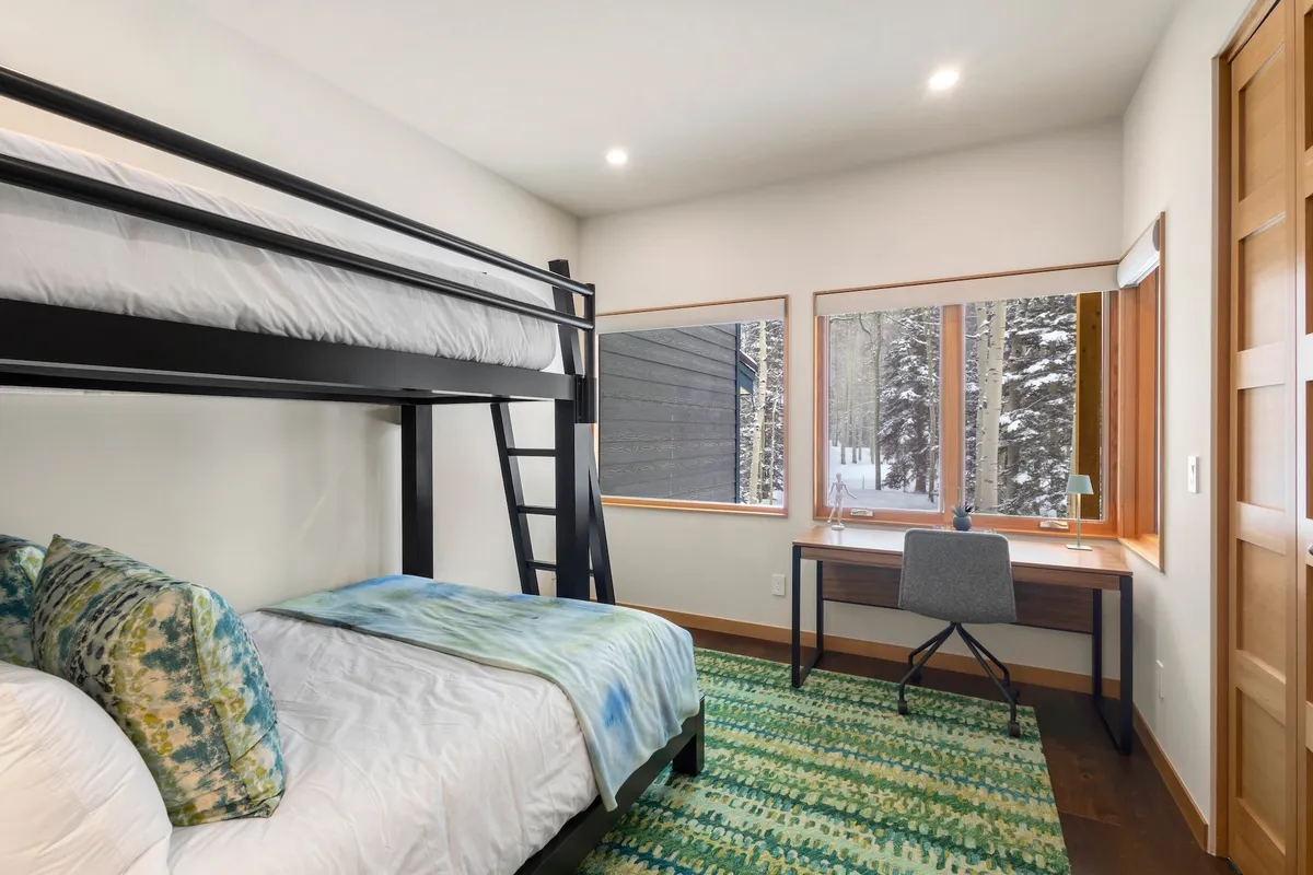 A guest room with a bunk bed, dedicated desk with a chair, and large windows letting in natural light. The space feels comfortable and serene, ideal for kids or guests to sleep, read, or work while enjoying snowy views outside.