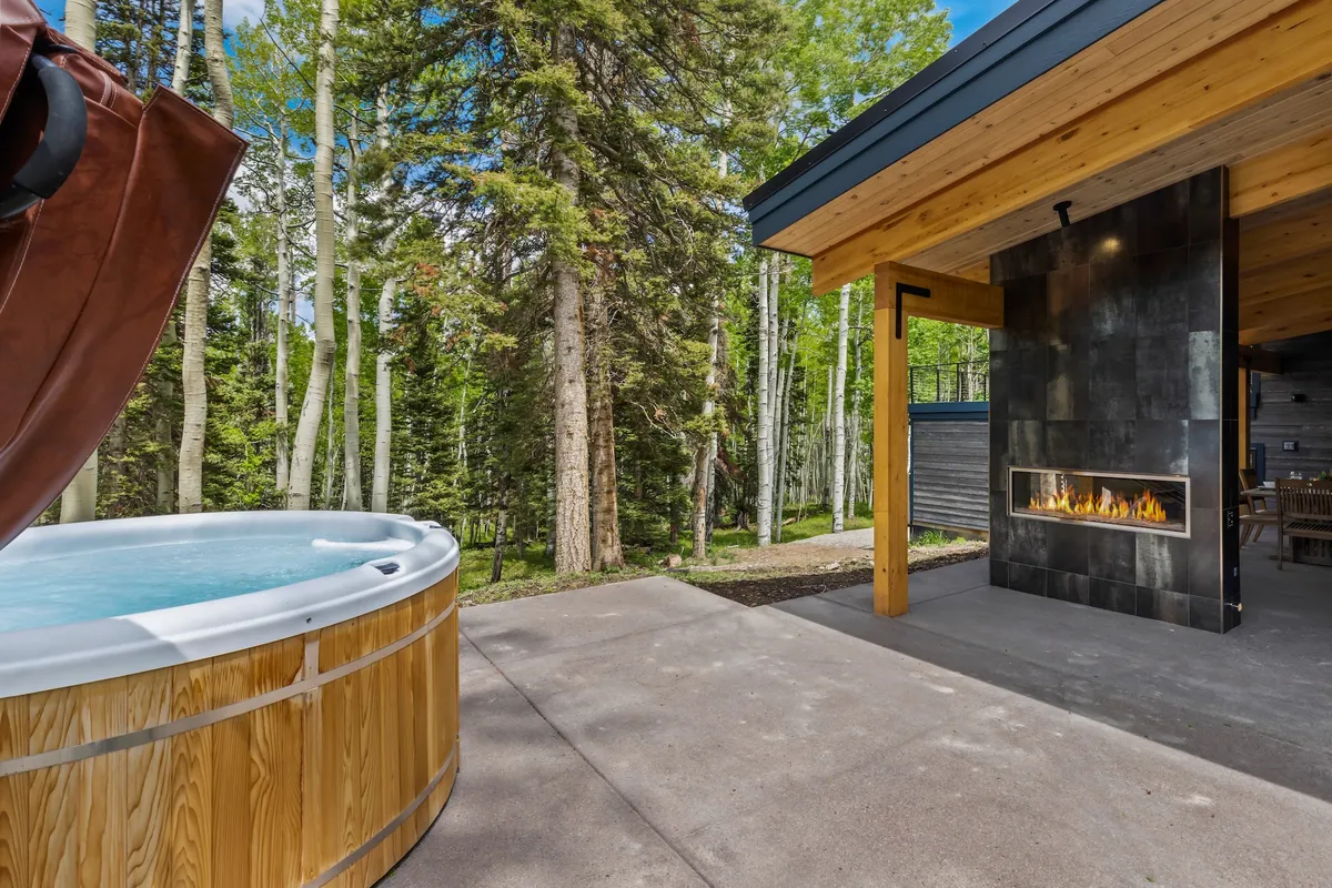 The photo shows a serene outdoor area with a hot tub and fireplace nestled among trees. The space invites relaxation, featuring comfortable seating and a backdrop of lush greenery, making it ideal for group conversations or stargazing after a day of activities.