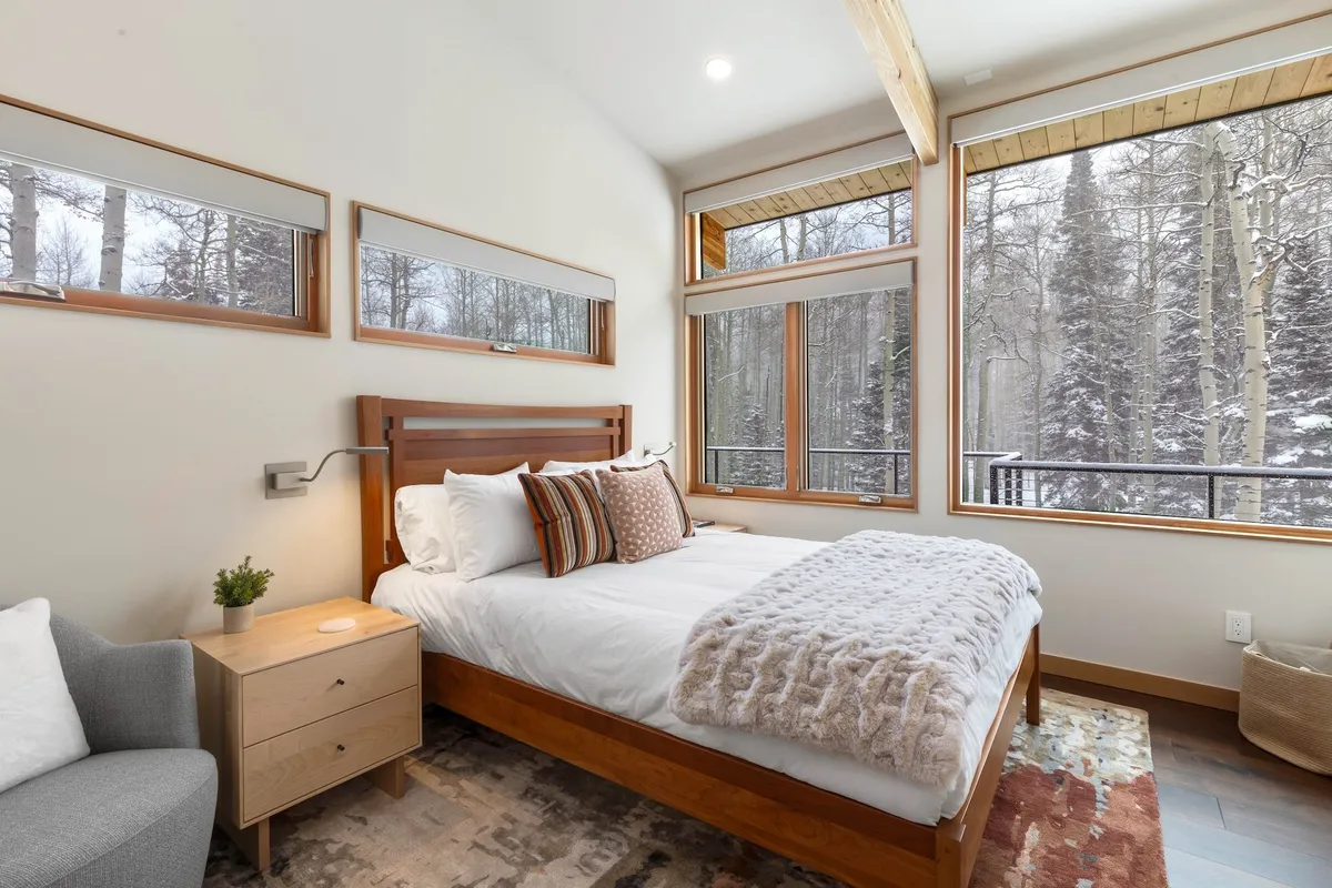 A plush bed with decorative pillows sits by large windows offering a sweeping forest view. A soft throw blanket and modern nightstand with a small plant create a cozy, inviting atmosphere perfect for relaxation and taking in the snow-covered landscape.