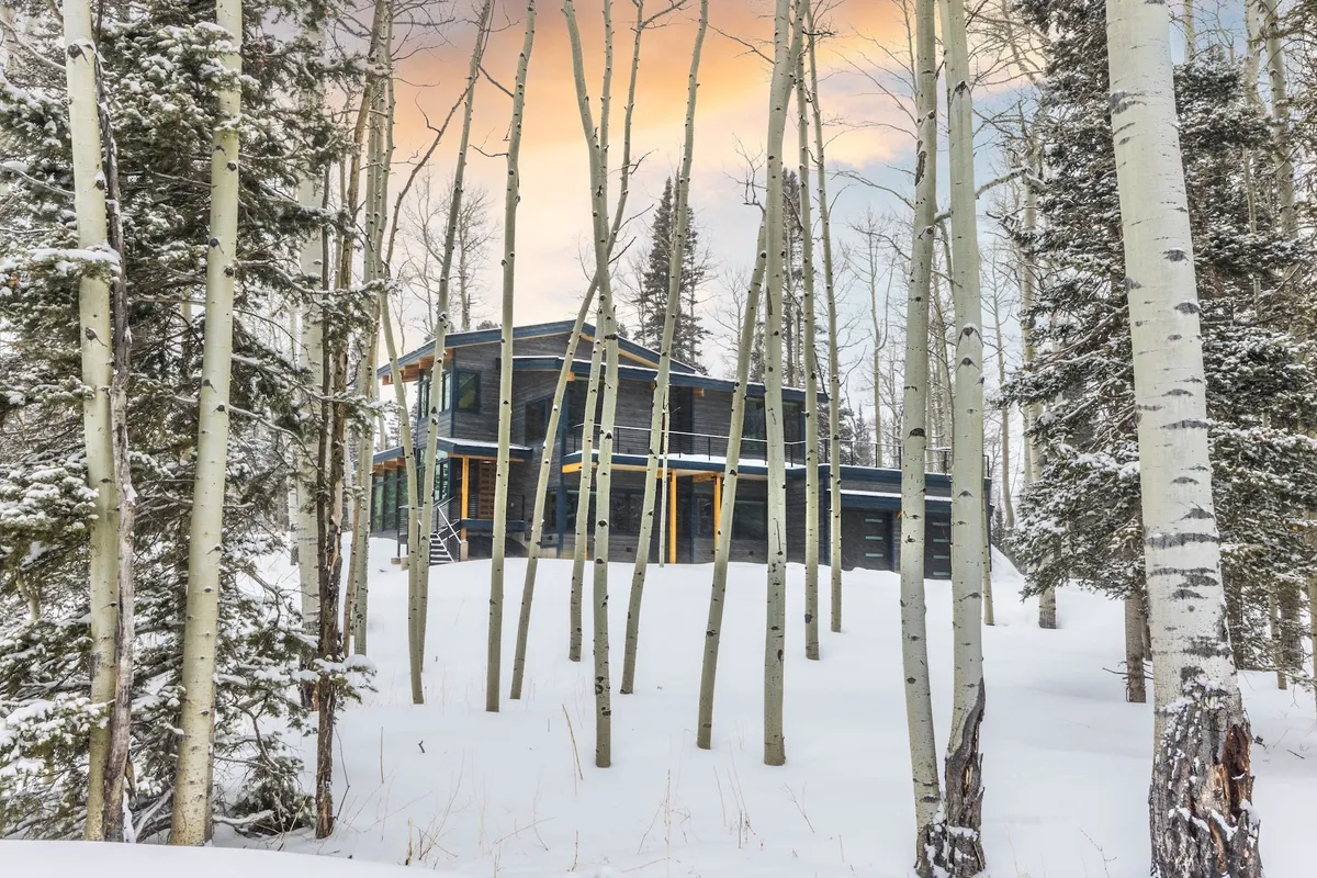 A stunning exterior view of the modern mountain home framed by aspen trees and a snow-covered landscape. The home appears both secluded and architecturally impressive, with soft evening light evoking a peaceful atmosphere.
