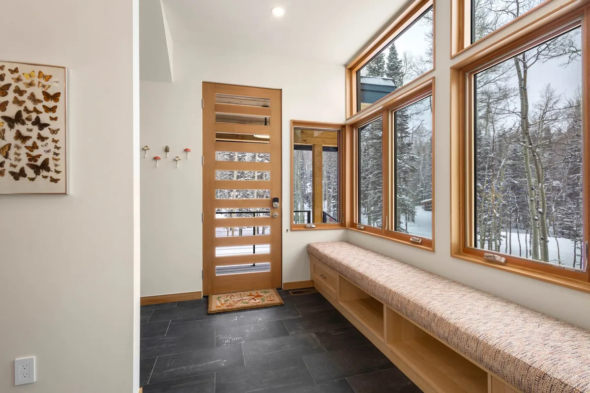 This image features a welcoming entryway with large windows that let in natural light, a sturdy wooden door, and benched seating. The space appears tidy and functional, with a serene view of snow outside, making it ideal for transitioning from outdoor adventures to the cozy indoors.