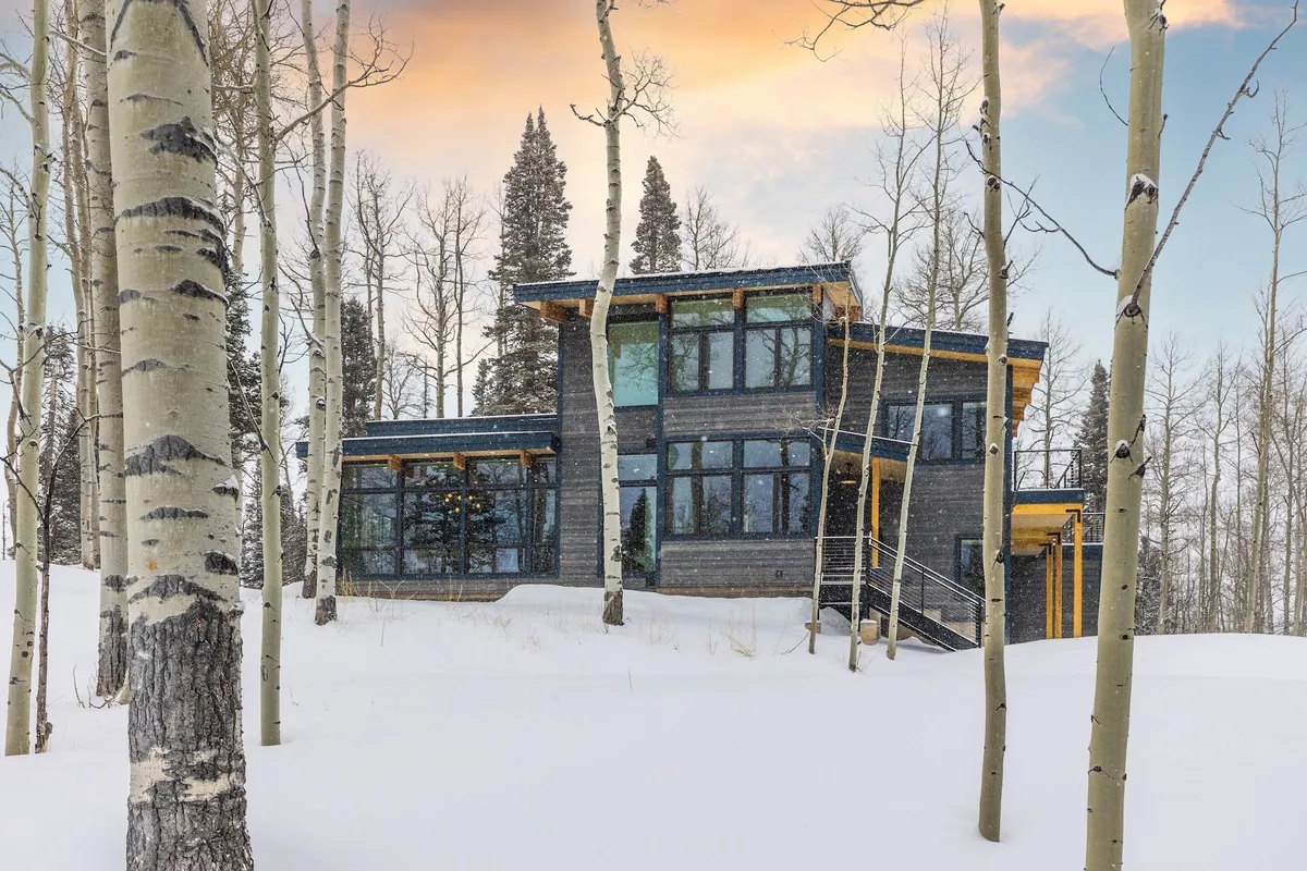 A modern house nestled among aspen trees, with a snow-covered yard and large glass windows that invite natural light. The scene is serene and family-friendly, hinting at winter activities like snow play or sledding in the spacious outdoor area.