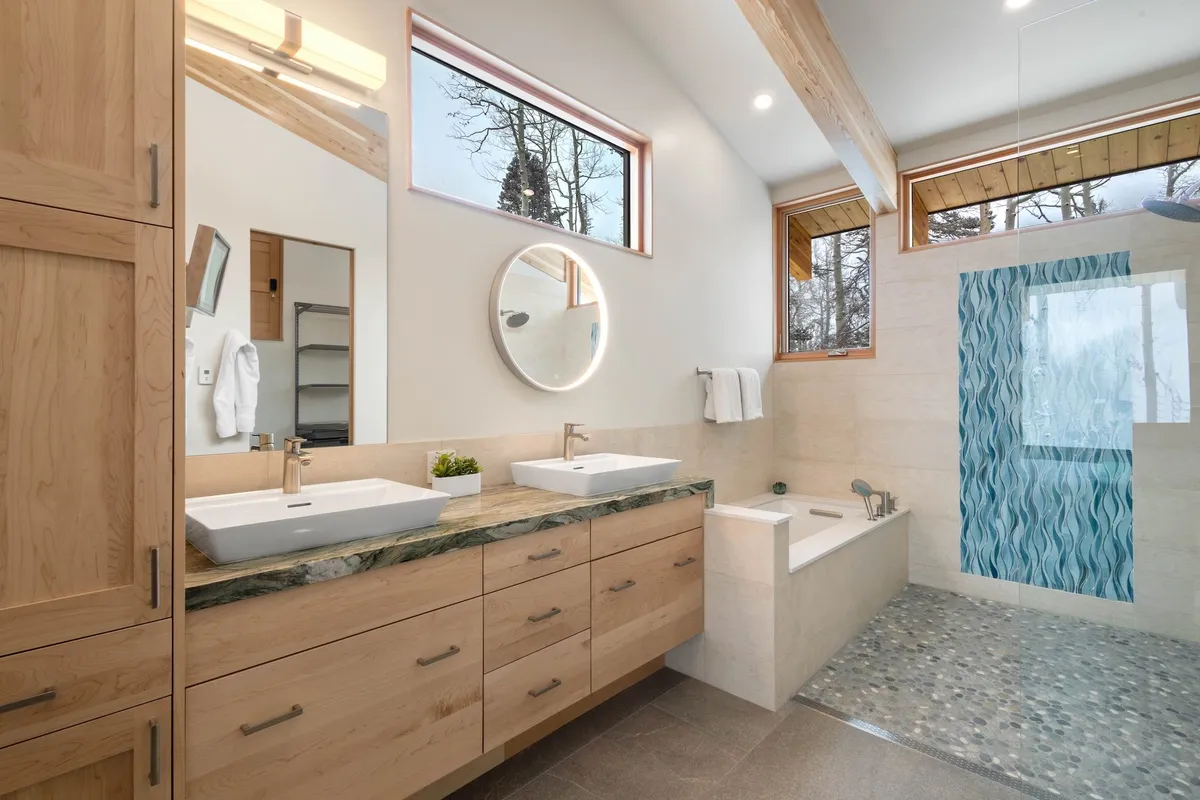 A spa-inspired bathroom with a double sink, circular mirror, blue wave-patterned shower wall, and abundant natural light streaming through windows. Exposed wooden beams add warmth and serenity to the space.