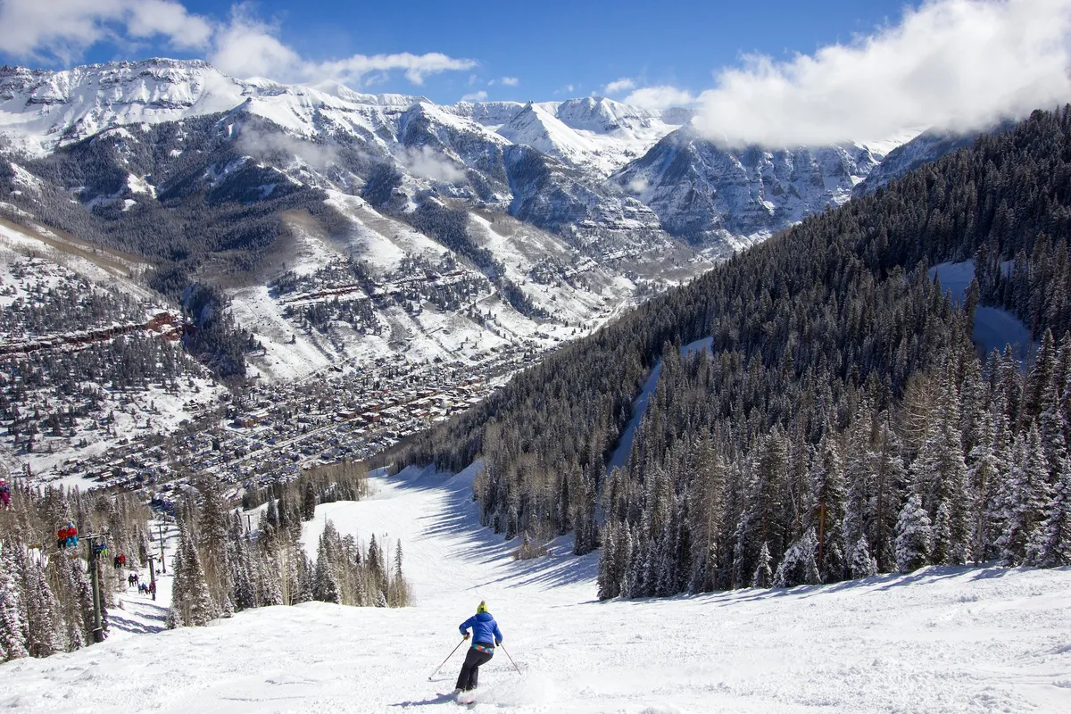 A skier glides down a snowy mountain slope with pine forests and the town visible below, encapsulating the thrill of nearby Telluride Ski Resort. The scene is dynamic, with adventure and natural beauty perfect for family ski days.