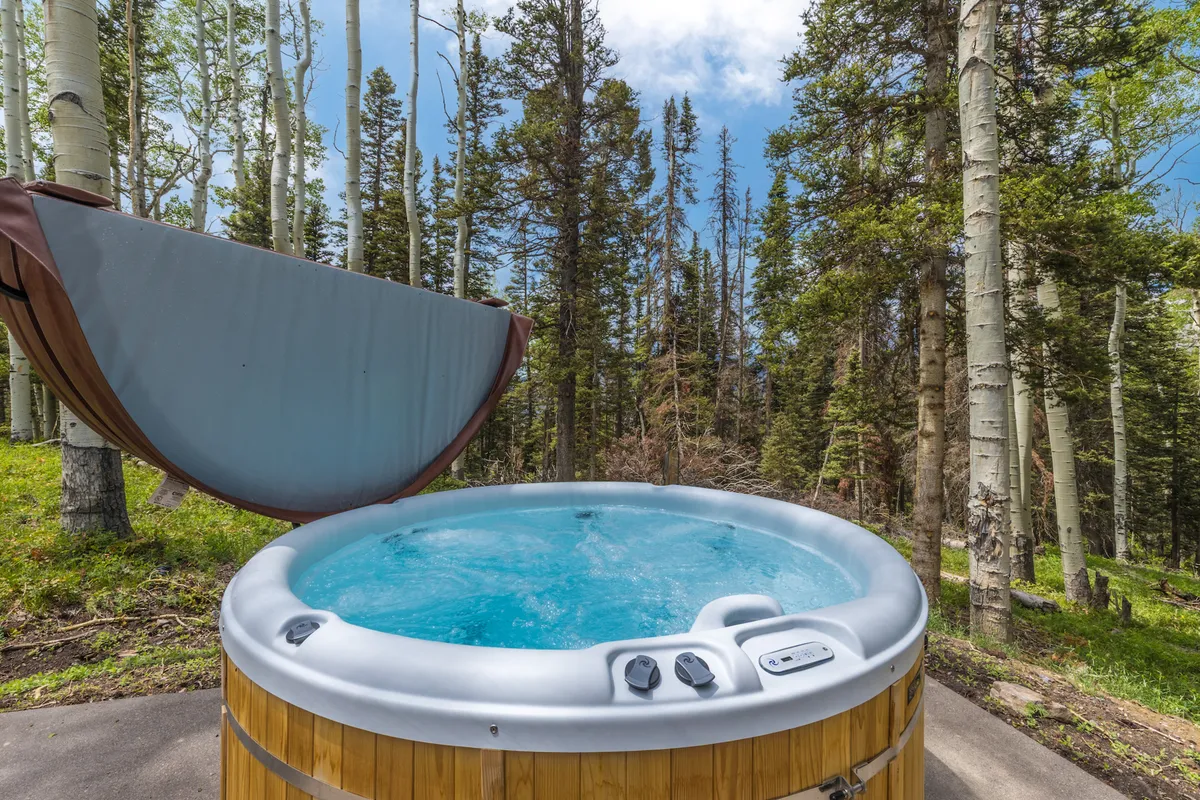 A serene outdoor hot tub nestled amidst tall trees with a clear blue sky overhead. The private hot tub area feels secluded and surrounded by nature, ideal for relaxation and intimate conversations.