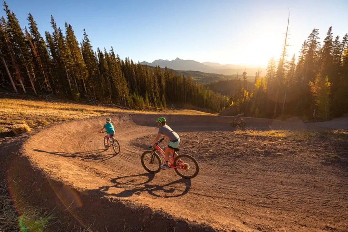 A group of mountain bikers rides along a curved dirt trail framed by pine trees and a dramatic mountain backdrop. The energy and movement highlight summertime mountain biking adventures, with the natural setting underscoring the area’s appeal for active recreation beyond winter sports.