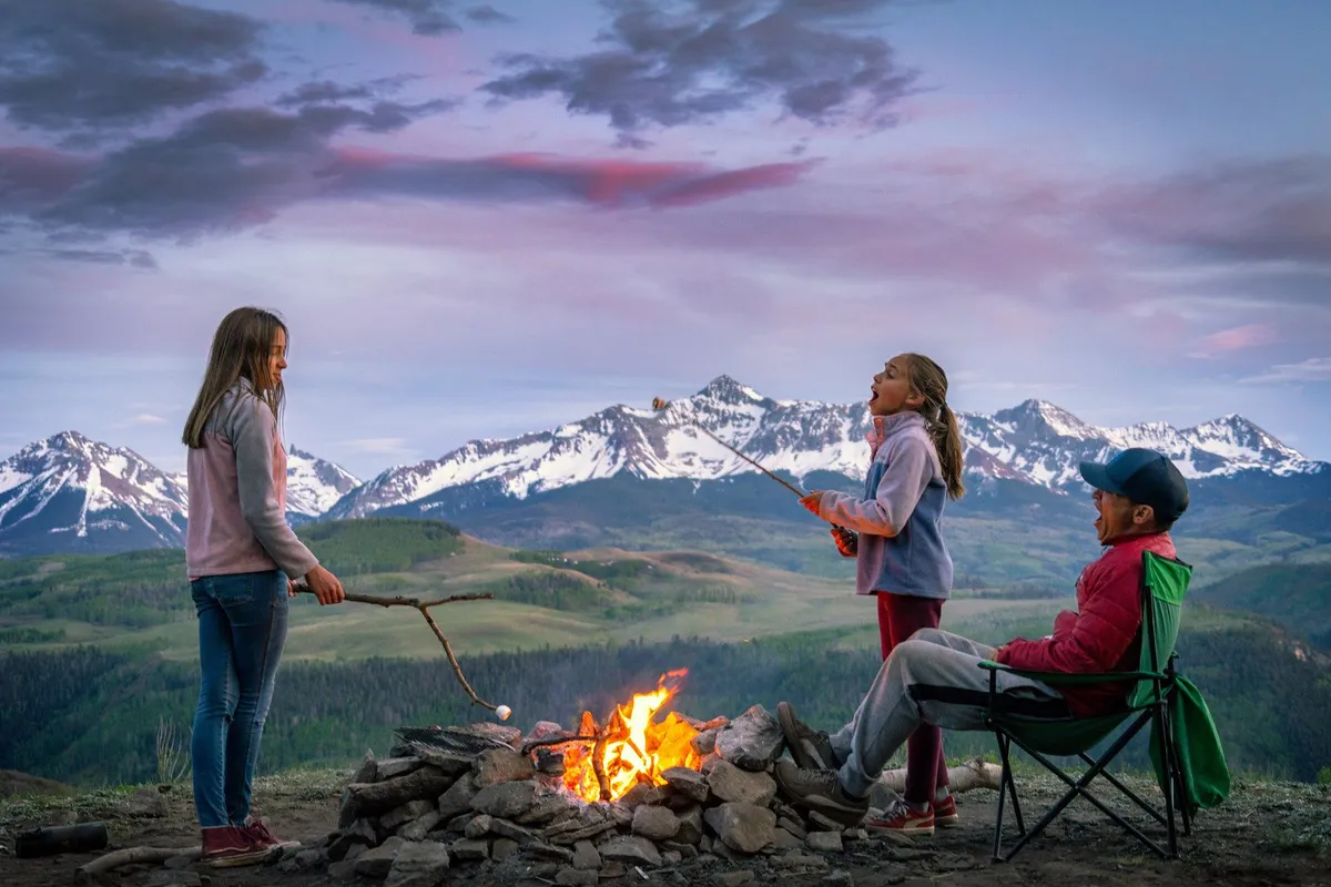 Children roast marshmallows over a campfire with a mountain backdrop, accompanied by an adult, evoking joyful, memorable family moments. The setting is outdoorsy and welcoming, ideal for year-round evening gatherings and storytelling.