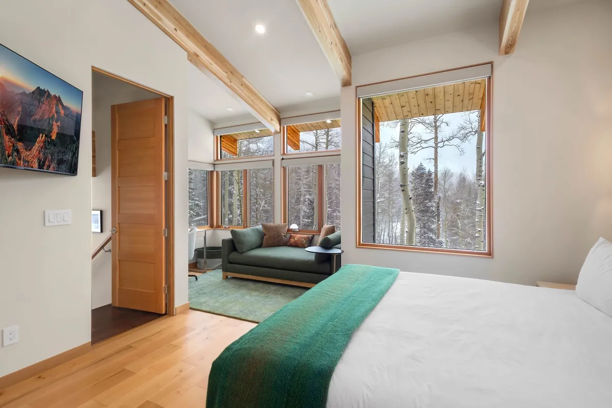 A serene and stylish guest suite with large windows revealing views of snow-covered trees, a modern sofa, a cozy bed with a green throw blanket, and a television mounted on the wall. The space feels calm and inviting, ideal for solo relaxation after lively group activities.