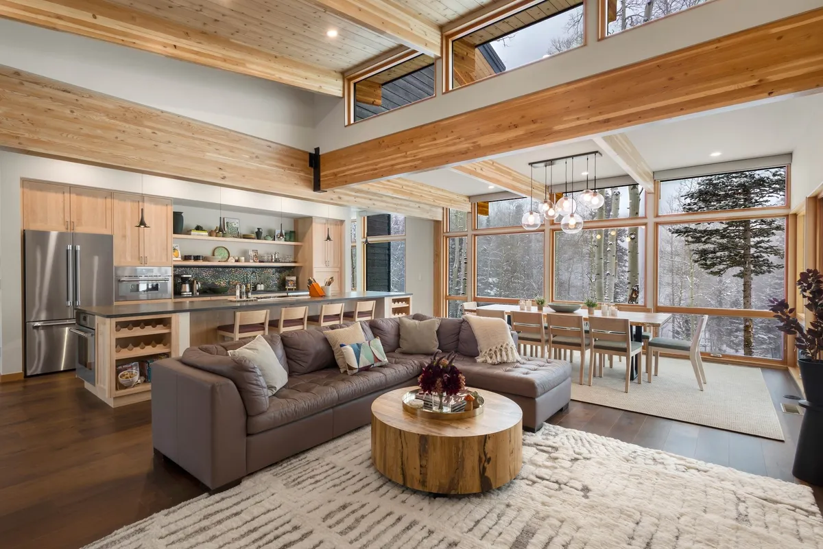 The hero image showcases a spacious, sunlit living area with expansive windows framing tranquil forest views, a plush sectional sofa perfect for gathering, a sleek wooden coffee table, and a modern dining area—all set against the backdrop of snowy Telluride landscapes.