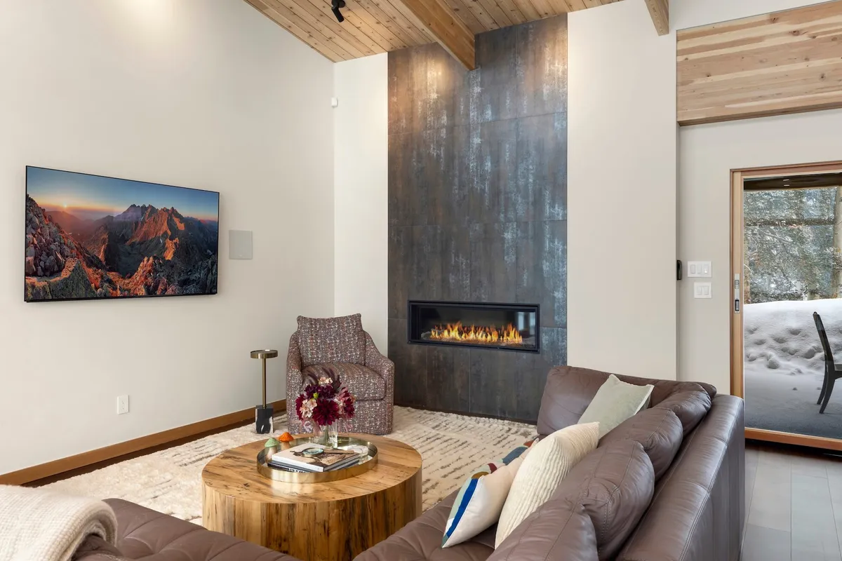 A warmly lit interior room featuring a modern fireplace, a stylish armchair, a large coffee table, and a flat-screen TV mounted on the wall. The windows offer a view of the outdoors, adding a sense of openness to the inviting space. The overall ambiance is ideal for relaxing by the fire, watching movies, or socializing with friends.