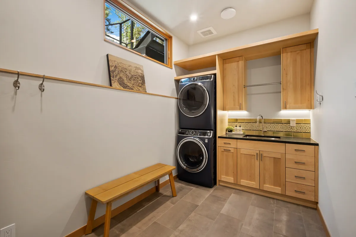 Depicts a practical mudroom/laundry area with a stacked washer and dryer, a deep sink with countertop, a wooden bench for changing or organizing, and decorative wall art. The space is both inviting and highly functional.