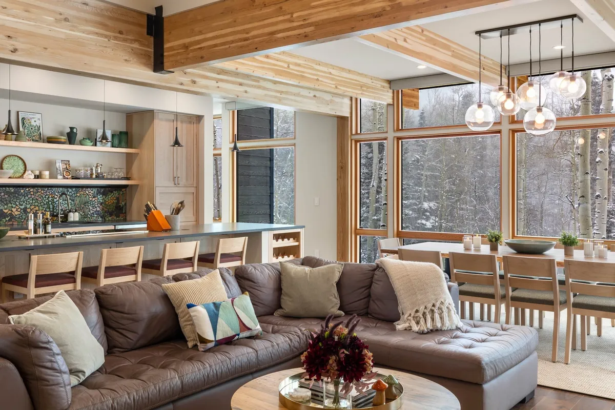 A warm, inviting open-plan living and dining area featuring large windows that frame snowy aspen trees outside. A comfortable sectional sofa and a spacious dining table sit under pendant lighting, with natural light pouring in, seamlessly connecting the interior with the surrounding landscape.