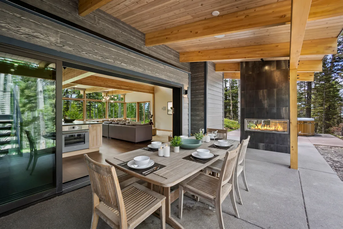 This image captures an outdoor dining area featuring a large table, a cozy fireplace, and expansive glass doors seamlessly connecting the indoor and outdoor spaces. The scene is framed by natural wooden beams and surrounded by trees, creating an inviting setting for gatherings and celebrations under the open sky.