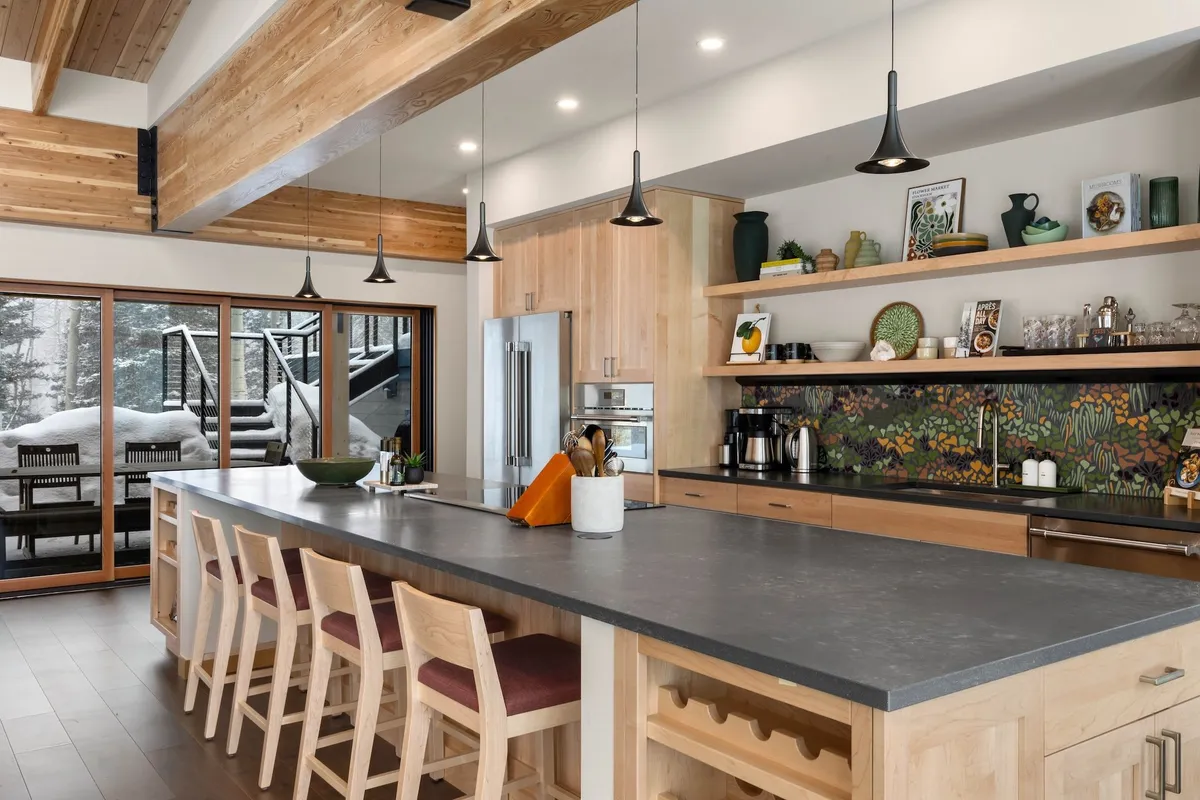 A modern gourmet kitchen with a large central island, stylish pendant lights, and a colorful backsplash. The image includes a glimpse of the outdoor deck, emphasizing a space designed for cooking, entertaining, and easy flow between indoors and out.