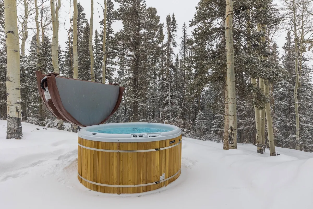 A steaming hot tub sits amidst a snow-covered forest, surrounded by trees. The inviting scene suggests a restorative soak after a day spent on the slopes or trails, with tranquil mountain ambience and cozy touches that evoke relaxation and recovery.